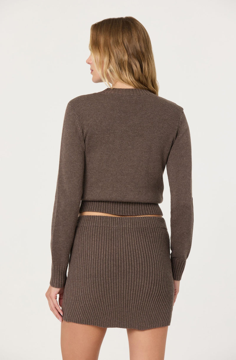 A woman is wearing a matching two-piece outfit made of brown ribbed knit fabric. The top is a long-sleeved, crewneck sweater that extends slightly above the waist. The skirt is a high-waisted, fitted mini skirt with vertical ribbing, ending mid-thigh.