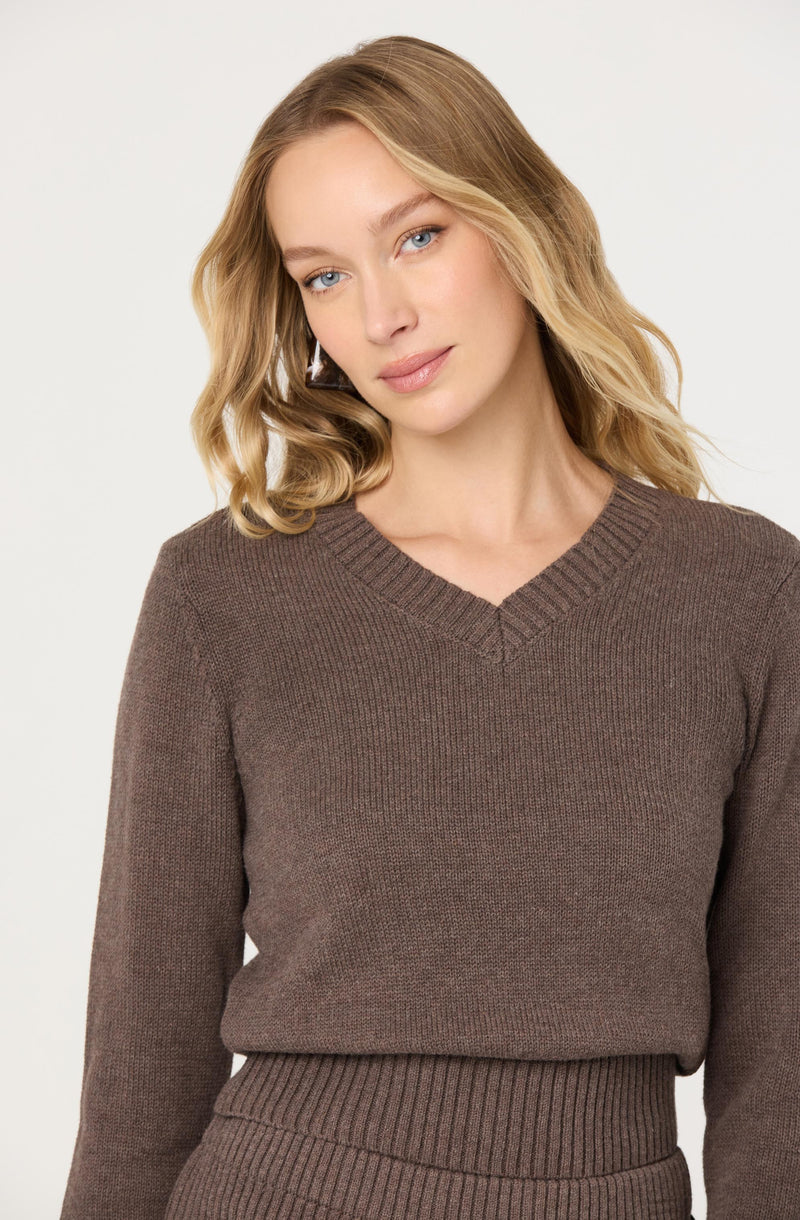 A woman is wearing a brown, long-sleeved, V-neck knit sweater with a ribbed neckline. She is also wearing matching ribbed knit pants.