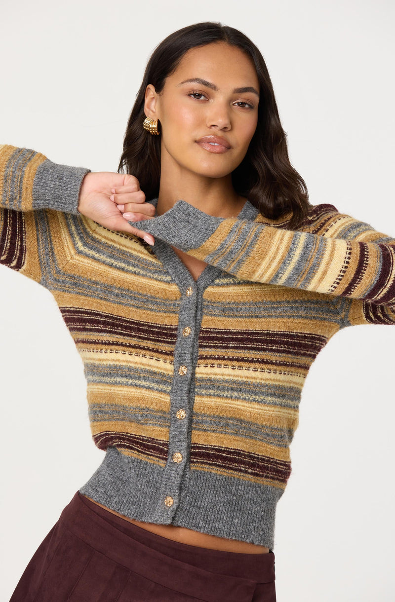 A woman is wearing a long-sleeved, button-up cardigan with horizontal stripes in shades of gray, yellow, brown, and maroon. The cardigan has a V-neckline, ribbed cuffs and hem, and features small, round, gold buttons down the front. She has dark, wavy hair, gold hoop earrings, and is posing with one arm raised and bent at the elbow, adjusting the collar of her cardigan. She has a neutral expression and is standing against a plain white background.