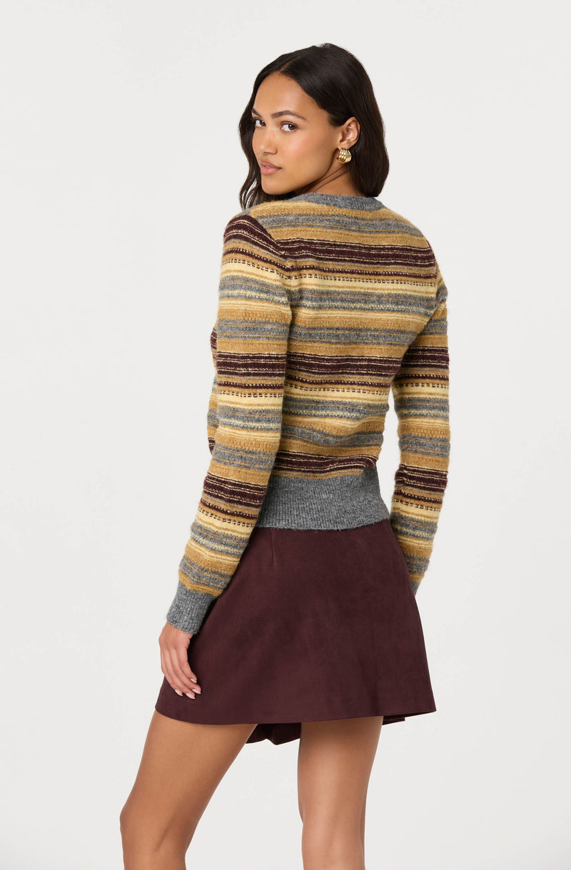 A woman is wearing a multicolored, striped sweater with shades of yellow, brown, gray, and rust, featuring a fitted gray waistband. She pairs it with a dark burgundy suede mini skirt.
