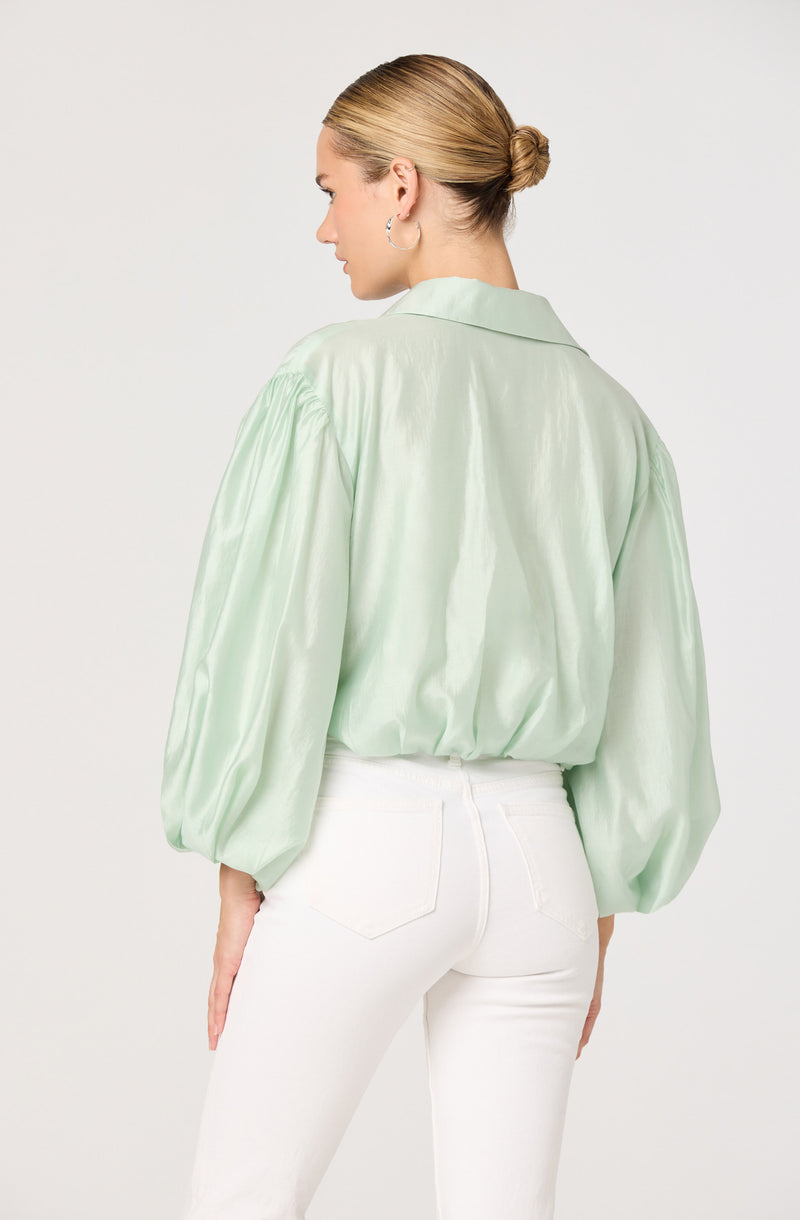 A woman with blonde hair tied in a bun is wearing a light green, satin-like blouse with long, puffed sleeves and a collar, paired with high-waisted white pants.