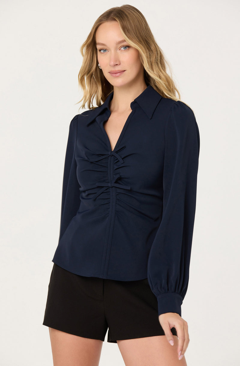 A woman is wearing a long-sleeved navy blue blouse with a collar and front ruching detail that creates a gathered effect at the center. The blouse has elasticized cuffs at the wrists. She is also wearing black shorts.