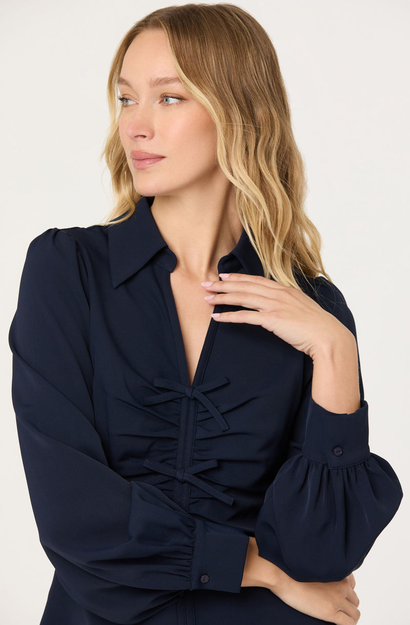 A woman wearing a navy blue button-down blouse with puffed long sleeves. The blouse features a front lace-up detail with ties.