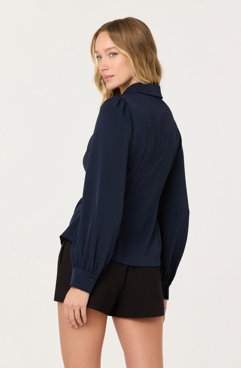 A woman is wearing a long-sleeved navy blue blouse with a collar and subtle pleats on the shoulders. The blouse has buttoned cuffs at the wrists. She pairs the blouse with black shorts.