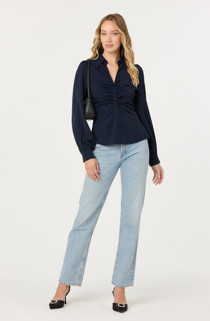 A woman is wearing a navy blue long-sleeve blouse with a collared neckline and a gathered, ruched design down the front. She pairs it with light-wash, straight-leg jeans and black high-heeled shoes with decorative embellishments. She carries a small black shoulder bag.