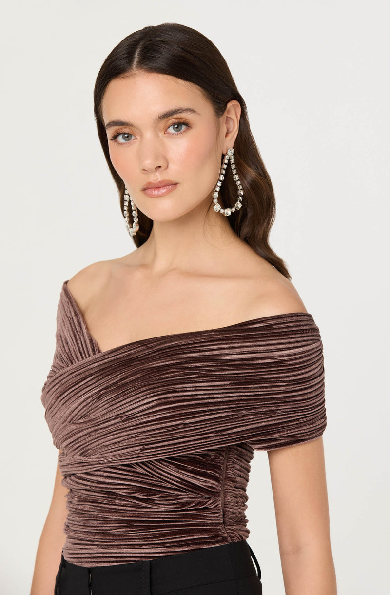 A woman with dark brown hair styled in soft waves is wearing large, silver hoop earrings adorned with sparkling stones. She has on an off-the-shoulder top in a textured, pleated fabric in shades of mauve and brown, which drapes across her shoulders and features horizontal pleats throughout. The top is fitted and gathered at the waist.