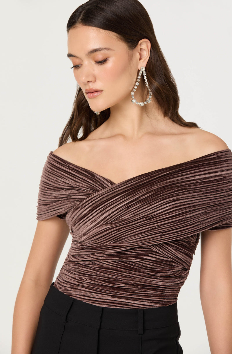 A woman with long, dark hair wearing large, sparkling hoop earrings with multiple clear stones. She is dressed in an off-the-shoulder top with a pleated, wrapped design in a mauve or dusty rose color. The top has a textured, shiny appearance and is fitted around the waist.
