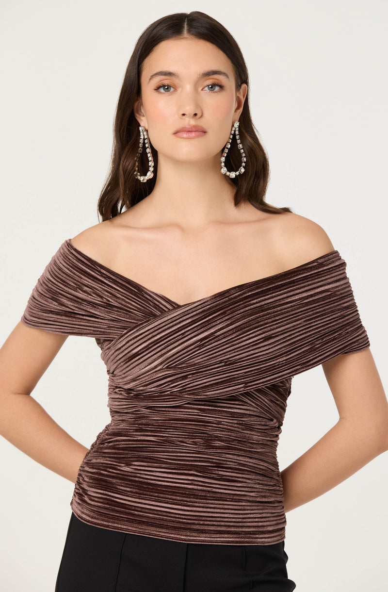 A woman is wearing an off-the-shoulder top made of shimmery, pleated fabric in a mauve or dusty rose color. The top has a ruched design that adds texture and dimension to the garment. She is also accessorized with large, dangling, rhinestone earrings.