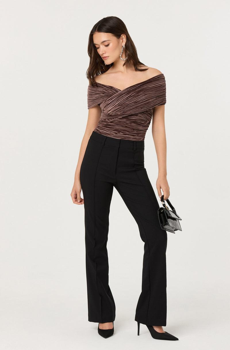 A woman is wearing an off-the-shoulder top with a pleated, textured fabric in shades of brown and mauve. She is paired with high-waisted, straight-leg black pants and black pointed-toe high heels. She is accessorizing with large, dangling earrings and is holding a small black handbag.