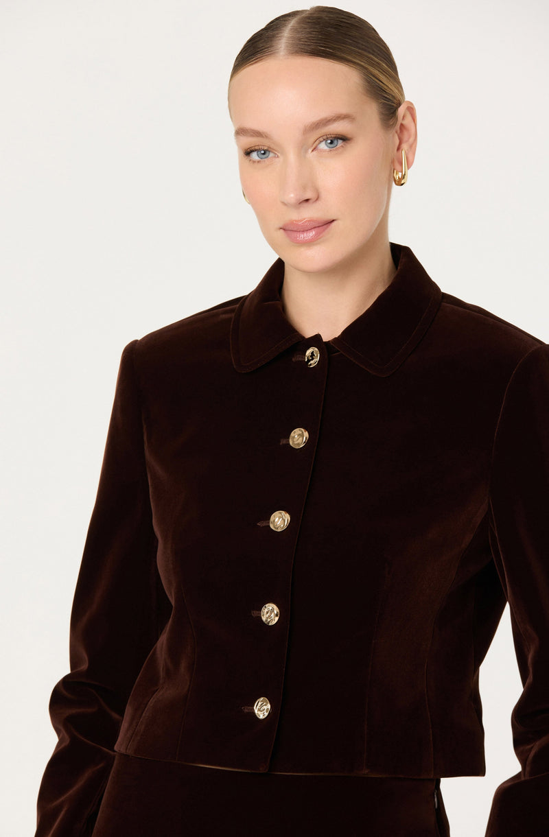 A woman is wearing a dark brown velvet cropped jacket with a classic collar and gold buttons down the front.