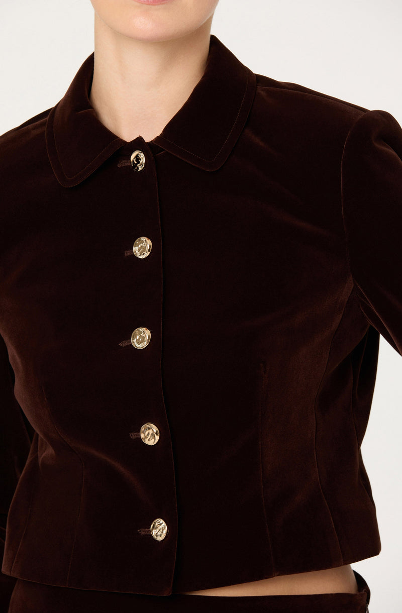 A woman is wearing a dark brown velvet button-up jacket with a classic collar. The jacket has shiny, round buttons down the front and features a fitted silhouette with seam detailing at the waist.