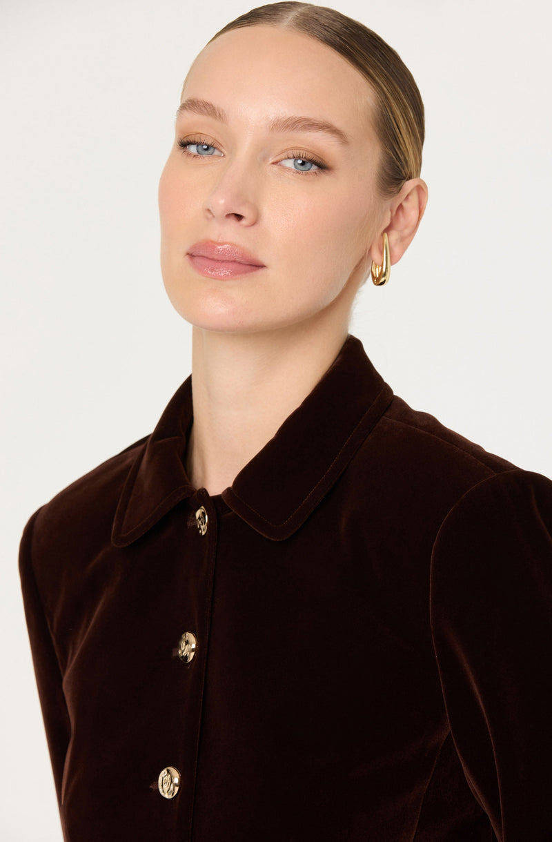 A woman with fair skin and blonde hair styled in a sleek bun is wearing a velvet shirt with a pointed collar and large shiny gold buttons down the front. She has small gold hoop earrings.
