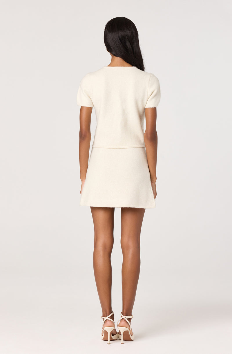 Woman wearing a cream short-sleeve top and matching skirt, paired with white high-heeled shoes.
