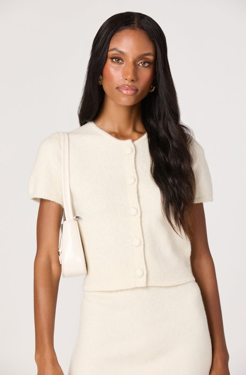 Woman in cream short-sleeve button-up sweater and matching skirt with a cream shoulder bag.