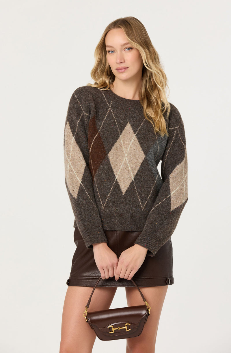 A woman is wearing a dark brown sweater with an argyle pattern in shades of beige, light brown, and red, featuring diamond shapes and diagonal lines. She is also wearing a dark brown leather skirt and is holding a small dark brown handbag with a gold-accented buckle.