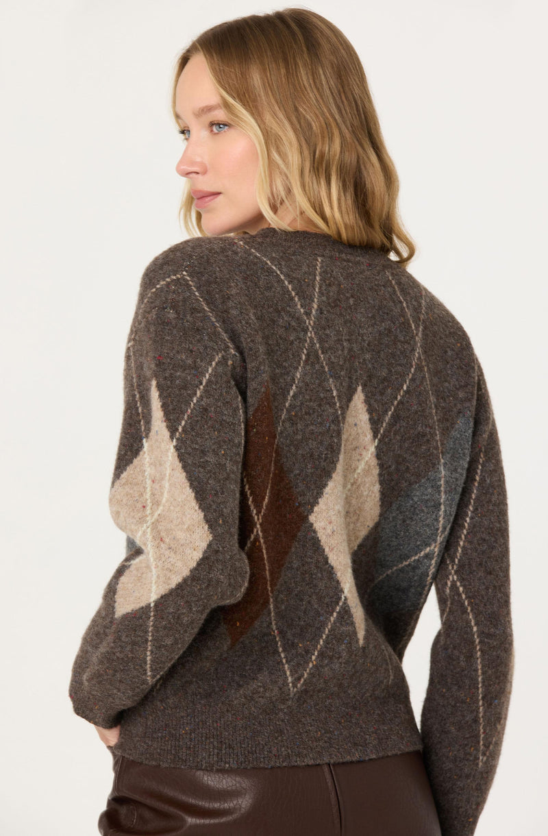 A woman is wearing a dark brown sweater with an argyle pattern featuring large diamond shapes in beige, light brown, and dark brown with fine white lines crisscrossing the diamonds. The sweater has a crew neckline and appears to be made of a soft, knit material. She is also wearing dark brown leather or faux leather pants.