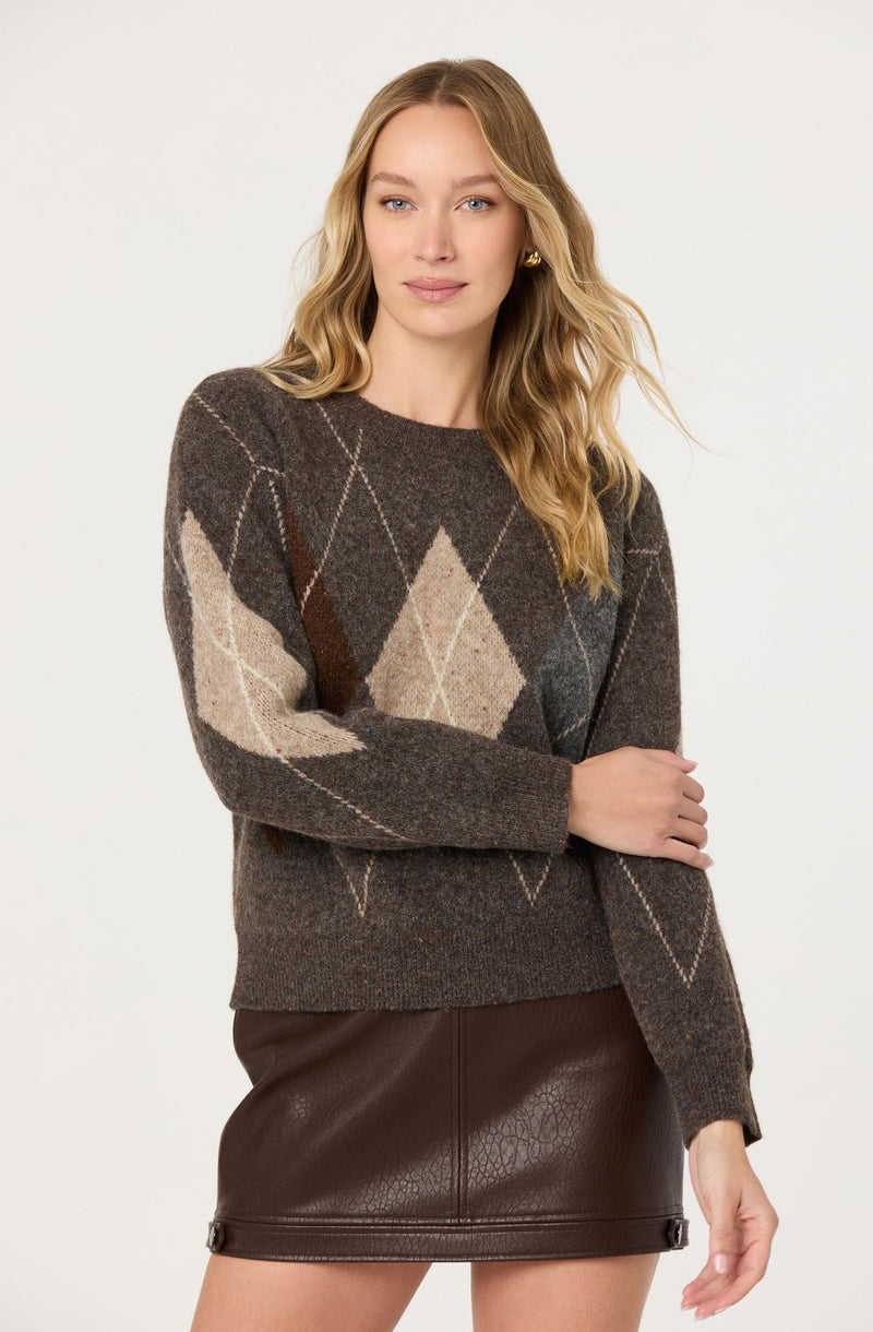 A woman is wearing a long-sleeve knit sweater with an argyle pattern in shades of brown, beige, cream, and dark gray. She is also wearing a dark brown leather skirt.