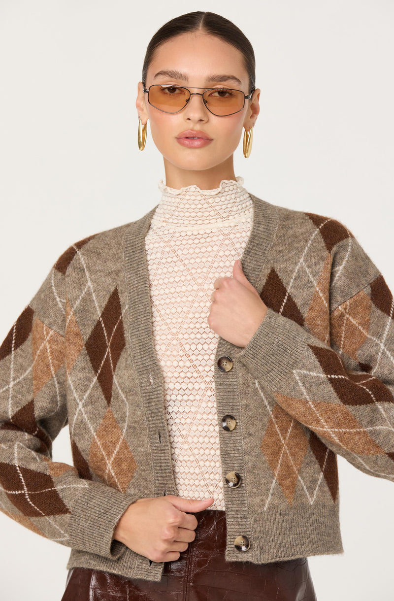 A woman is wearing a beige and brown argyle patterned cardigan with large tan and darker brown diamonds outlined in white. Underneath, she has on a high-neck white lace blouse. She accessorizes with gold hoop earrings and tinted sunglasses.