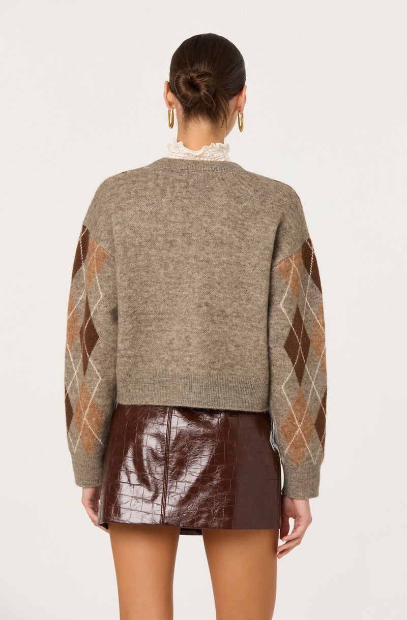 A woman is wearing a beige knit sweater with an argyle pattern in shades of brown, tan, and white on the sleeves. She has paired it with a shiny brown leather mini skirt. She is also wearing large gold hoop earrings and has her hair styled in a neat bun. Additionally, she is wearing a white lace high-neck top underneath the sweater.