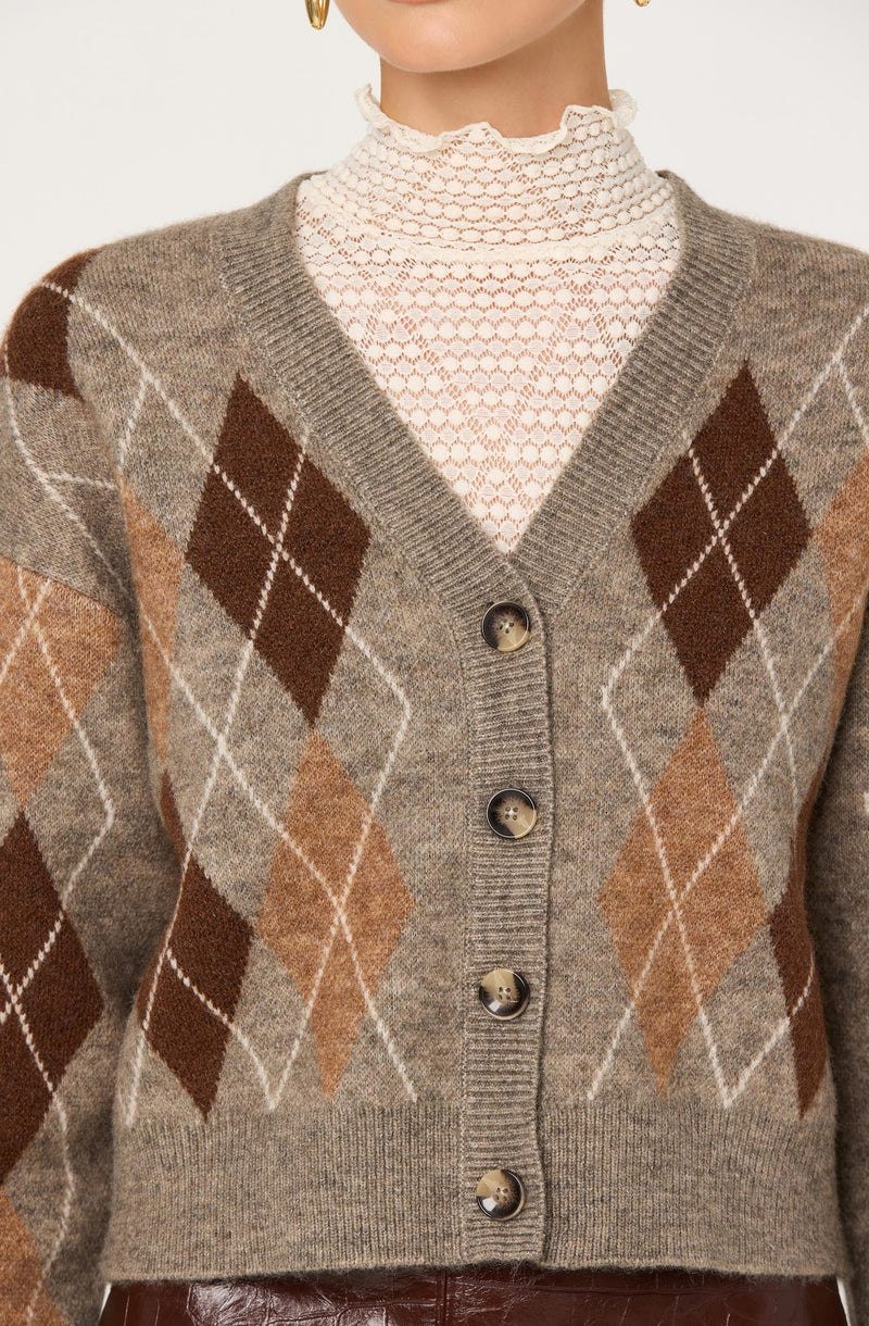 A person is wearing a brown, beige, and tan argyle patterned cardigan with a V-neck and large buttons. Underneath, they have on a high-neck, white lace or crochet top with a ruffled collar.
