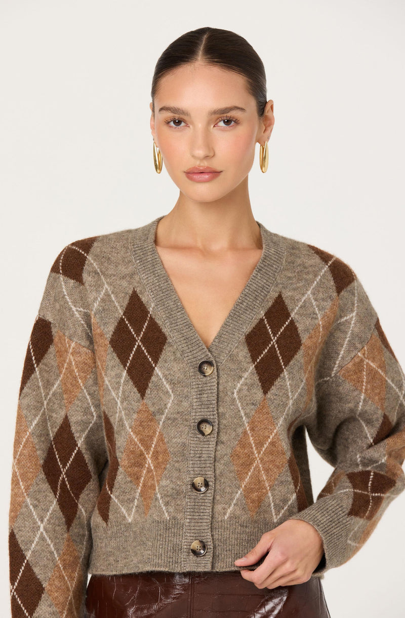 A woman is wearing a knitted cardigan with an argyle pattern featuring large diamonds in shades of brown, tan, and beige, with intersecting lines in white. The cardigan has a V-neckline and is fastened with four buttons down the front. She is also wearing large gold hoop earrings.