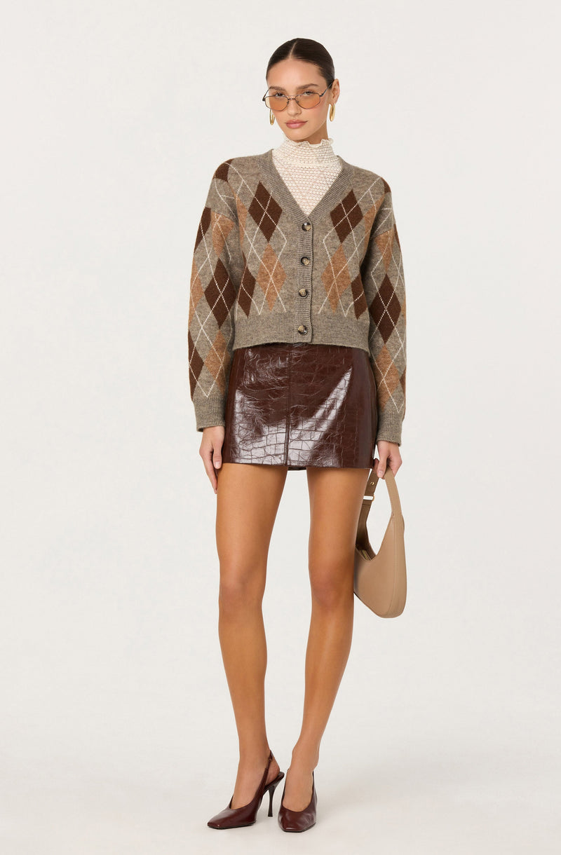 A woman is wearing a beige cardigan with an argyle pattern in shades of brown, tan, and gray over a white high-neck blouse with a subtle pattern. She has on a shiny, dark brown leather mini skirt and matching dark brown high heels. She accessorizes with gold hoop earrings, small glasses, and holds a beige handbag.