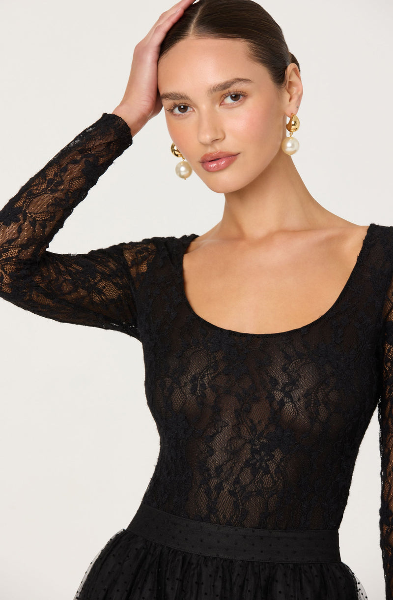 A woman is wearing a black lace long-sleeve top with a scoop neckline. The top is semi-sheer, revealing her skin underneath, and features a floral lace pattern. She is also wearing large pearl earrings with gold accents.
