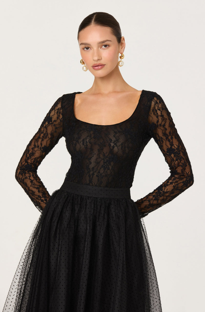 A woman is wearing a black lace long-sleeve top with a scoop neckline. The top is semi-sheer with a floral lace pattern. She is also wearing a black, high-waisted skirt made of a tulle fabric with small polka dots, which has a gathered waistline. She has large pearl and gold earrings.