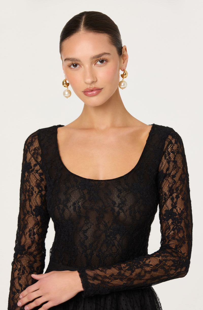 A woman is wearing a fitted black lace long-sleeve top with a scoop neckline. She accessorizes with large gold hoop earrings that have a pearl detail hanging from each hoop.