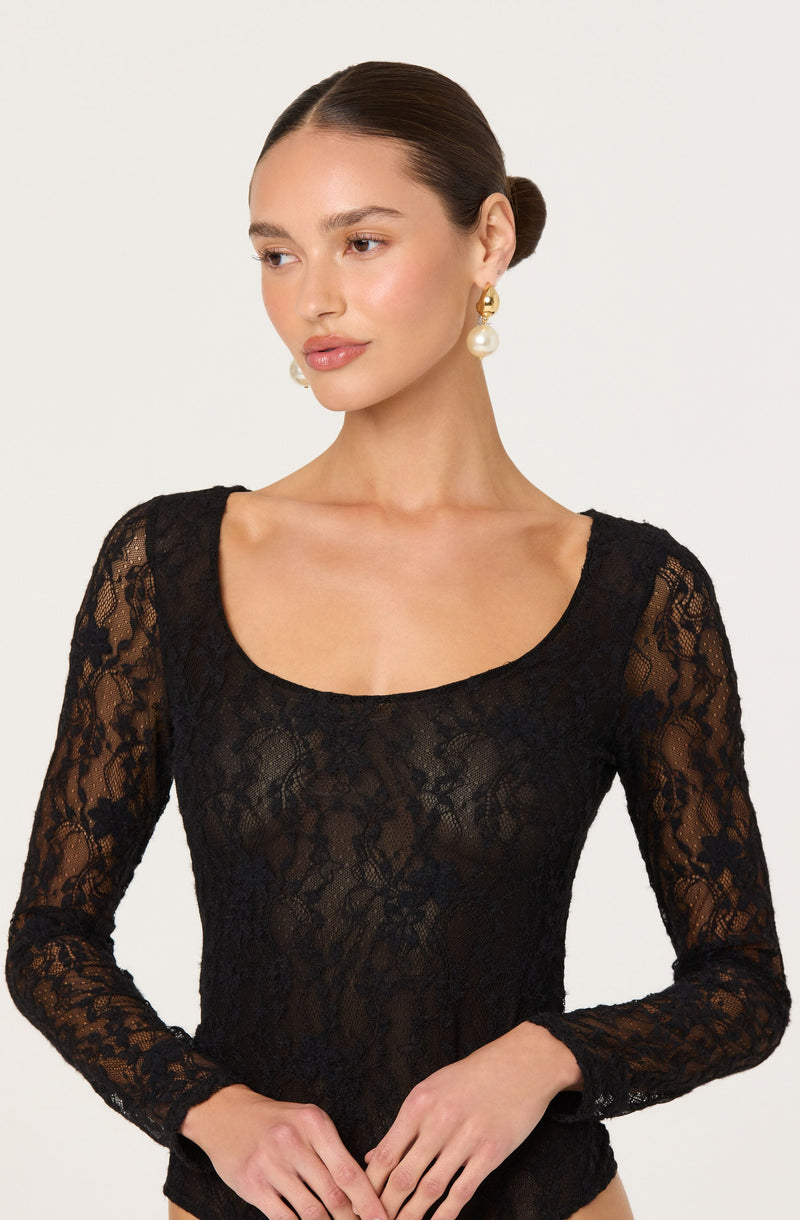A woman is wearing a black lace long-sleeve top with a wide, rounded neckline. The lace fabric features a floral pattern, and the top is sheer, revealing a hint of skin underneath. She is also wearing large, gold hoop earrings with a prominent pearl pendant hanging from each hoop.