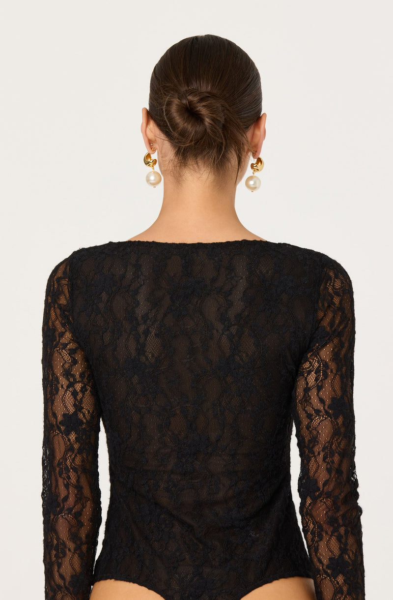 A woman is wearing a black lace long-sleeve top. The top is sheer with an intricate floral lace pattern. She has her hair styled in a neat bun and is accessorized with large, gold, and pearl drop earrings.