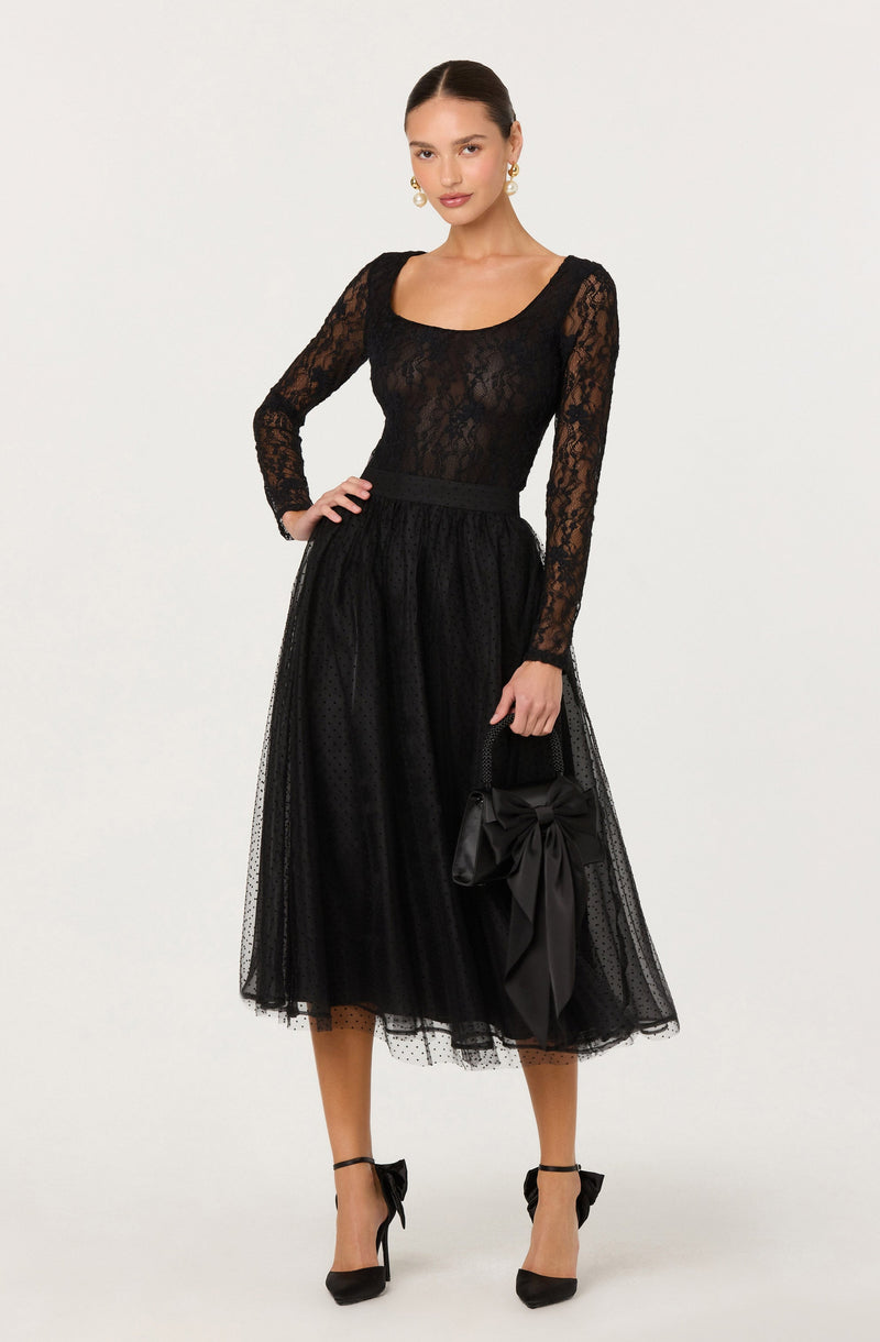 A woman is wearing a black lace long-sleeve top with a scoop neckline, paired with a black tulle midi skirt that has a subtle polka dot pattern. She accessorizes with large pearl earrings, black high-heeled shoes with ankle straps and bows, and holds a black handbag adorned with a large satin bow.