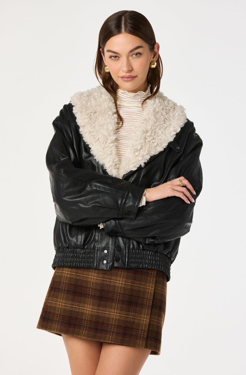 Woman wearing a black leather jacket with a cream shearling collar, plaid skirt, and gold earrings.