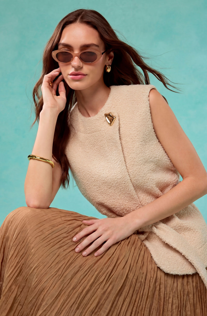 Woman wearing beige textured sleeveless top with a gold brooch and sunglasses, and a pleated brown skirt.