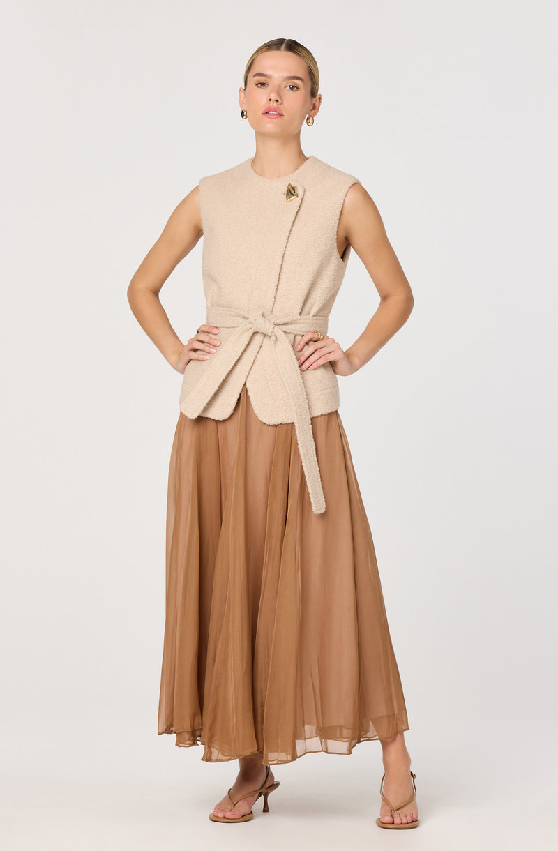 The outfit consists of a sleeveless, beige, textured vest with a decorative pin on the left shoulder and a matching waist belt tied in a bow. The vest is paired with a long, flowing, brown tulle skirt that reaches the floor. The model is also wearing light pink, open-toed high heels.
