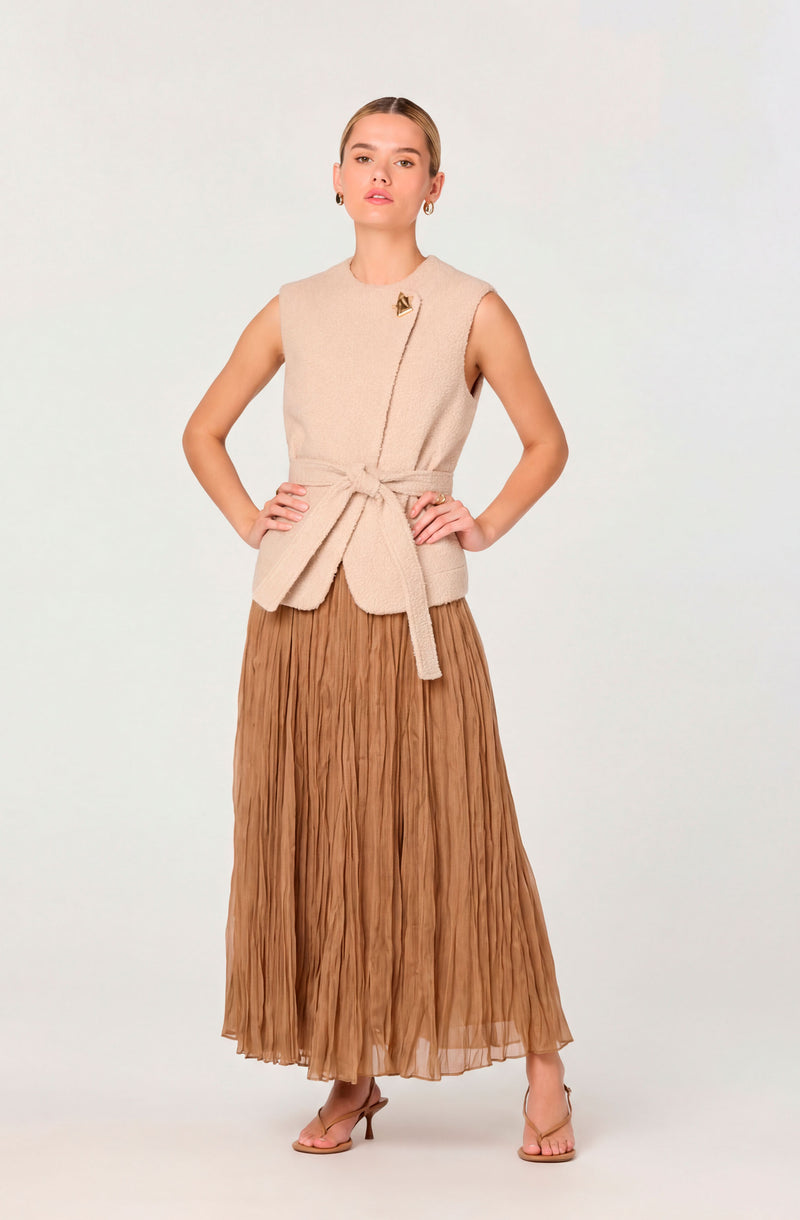 Woman in beige sleeveless vest with a belt and a gold pin, wearing a brown pleated maxi skirt and heels.