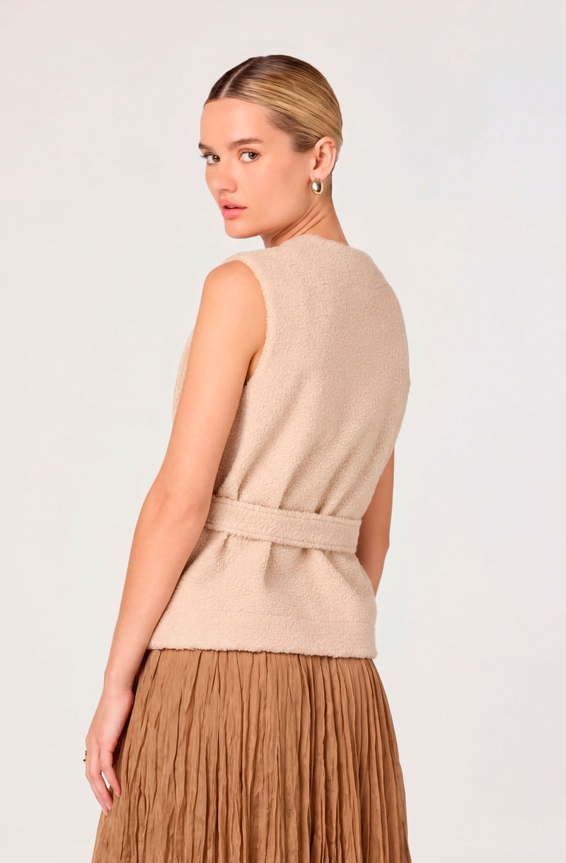 Woman wearing a sleeveless beige textured top with a belt and a brown pleated skirt.