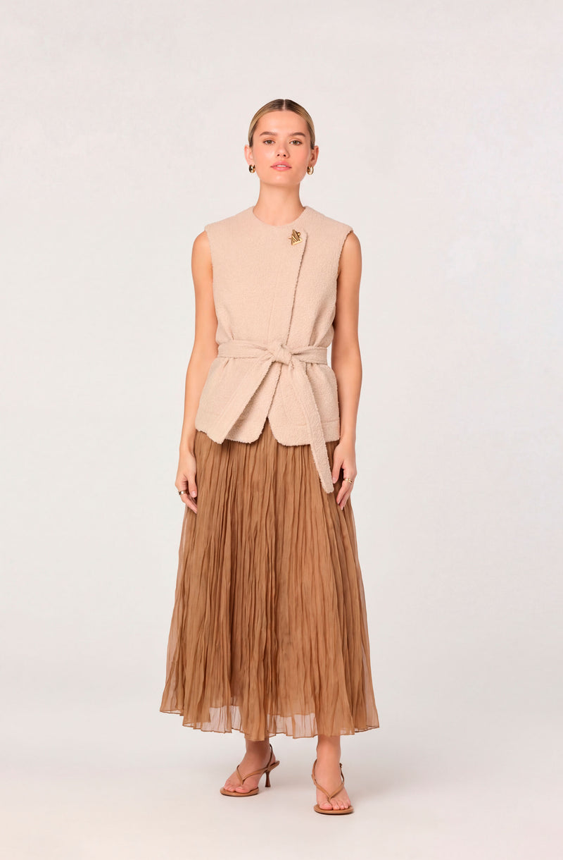 Woman wearing a sleeveless beige knit vest with a tied waist, paired with a brown pleated maxi skirt and nude heels.