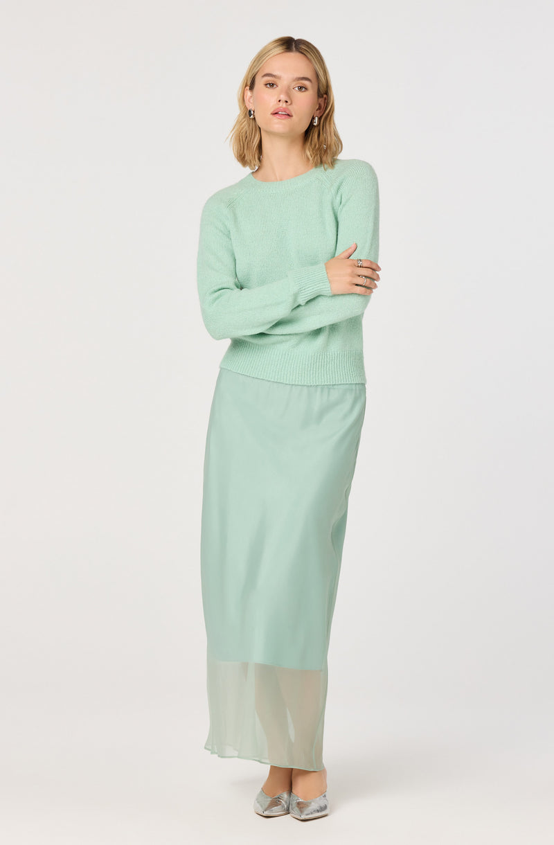 The woman is wearing a light mint green sweater with long sleeves and a crew neckline, paired with a matching long, sheer, flowy skirt. She has silver pointed-toe shoes and is accessorized with earrings and rings.
