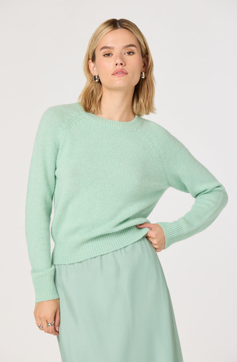 A woman is wearing a light mint green knit sweater with a round neckline and long sleeves. The sweater has a slightly loose fit and features a ribbed hem and cuffs. She is also dressed in a matching mint green skirt that appears to be made of a smooth, flowing fabric. She has shoulder-length blonde hair and is accessorizing with hoop earrings.