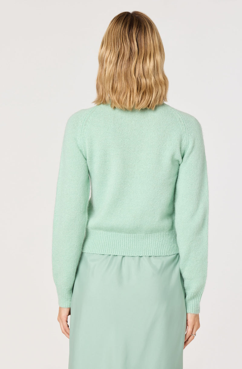 A woman is wearing a light mint green sweater with long sleeves and a ribbed hem and cuffs. She is also wearing a matching mint green skirt. The sweater appears soft and slightly fuzzy in texture. The outfit is monochromatic in soft pastel shades.