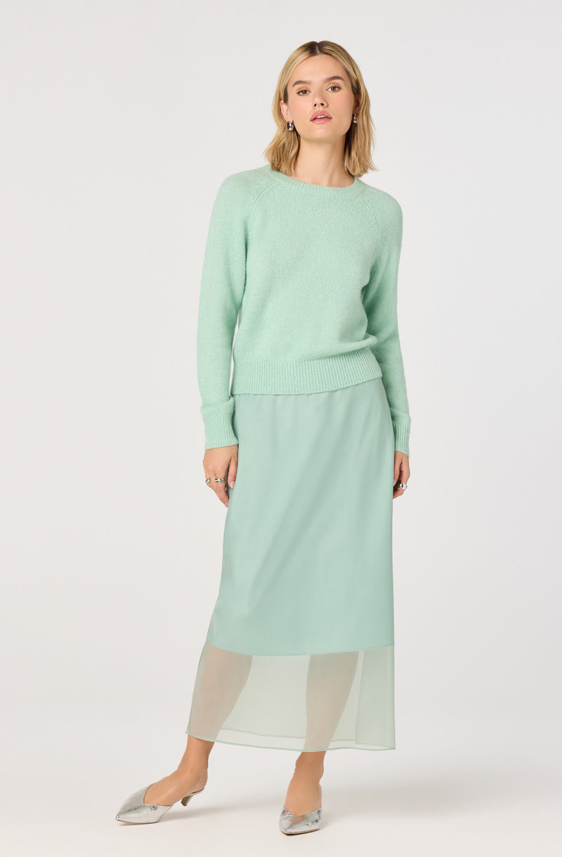 A woman is wearing a light mint green, long-sleeved knit sweater with a round neckline and ribbed cuffs, paired with a matching pastel green, semi-sheer, ankle-length skirt. The skirt has a solid interior layer ending at mid-calf, with a transparent overlay extending to the ankles. She is also wearing silver pointed-toe flats.