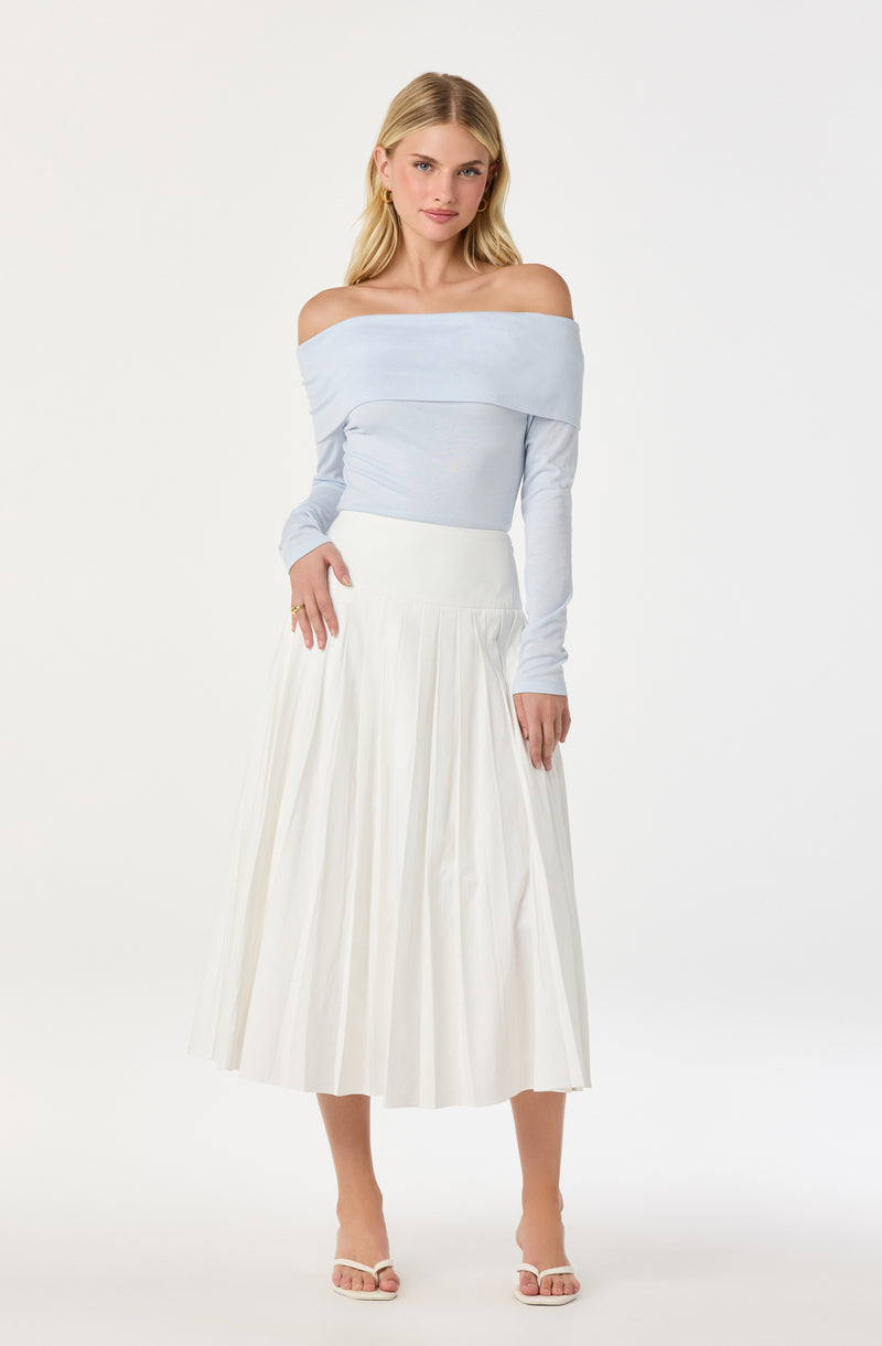 A woman is wearing an off-the-shoulder light blue long-sleeve top and a white pleated midi skirt. She has blonde hair, is accessorized with small hoop earrings, and is wearing open-toe heels.