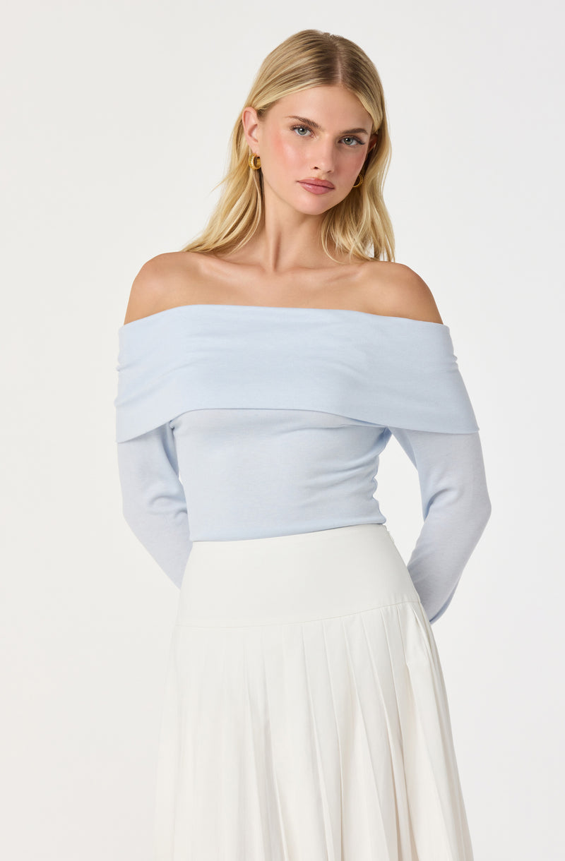 A woman is wearing an off-the-shoulder light blue top with a wide, fold-over neckline that drapes across her shoulders. The top has long sleeves and a fitted silhouette. She pairs it with a high-waisted, long white skirt that features soft pleats. She accessorizes with gold hoop earrings. The background is plain white.
