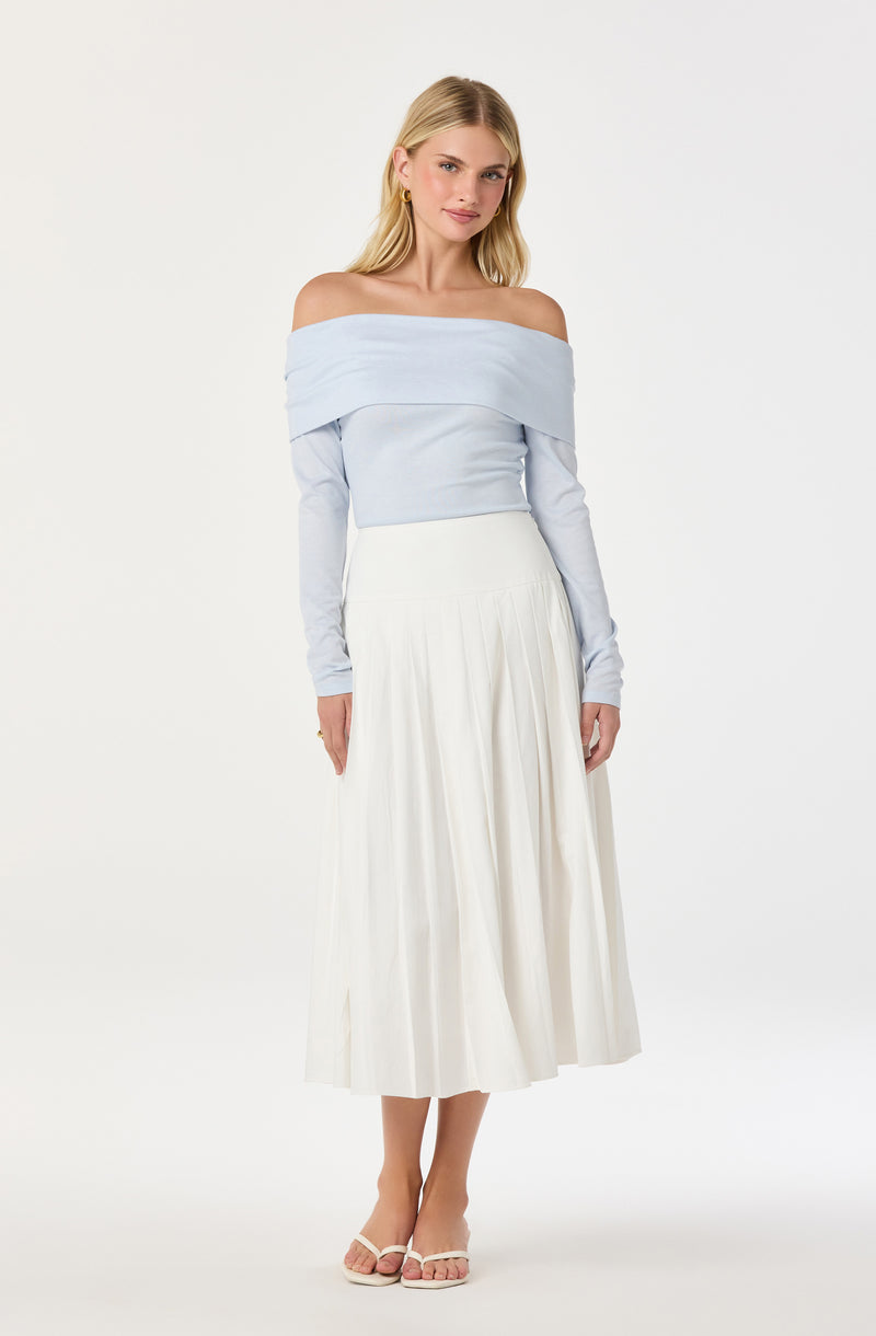 A woman is wearing an off-the-shoulder light blue top with long sleeves, paired with a high-waisted, white pleated midi skirt. She has blonde hair and is accessorized with gold hoop earrings and a gold ring. She is also wearing light-colored, open-toe heeled sandals.