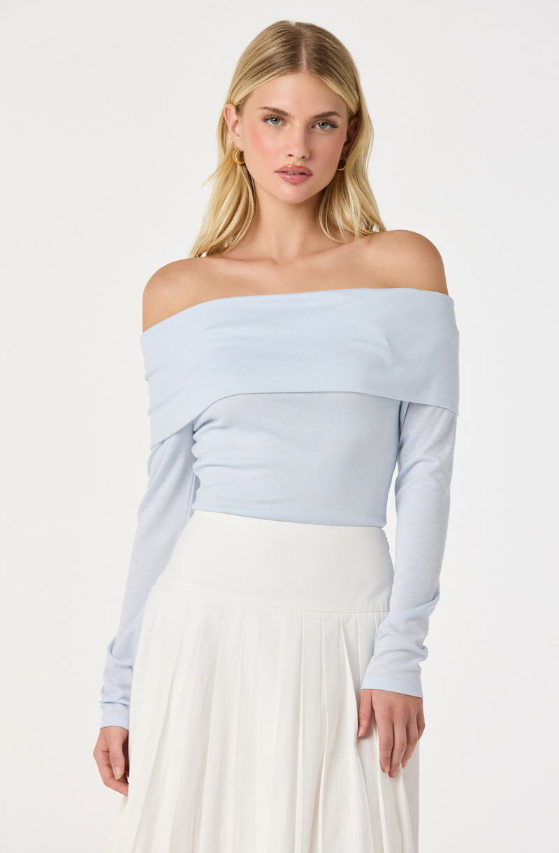 The woman is wearing an off-the-shoulder light blue top with long sleeves and a wide neckline that drapes over her shoulders. She pairs it with a high-waisted, flowing white skirt that has pleats. She also has on gold hoop earrings.