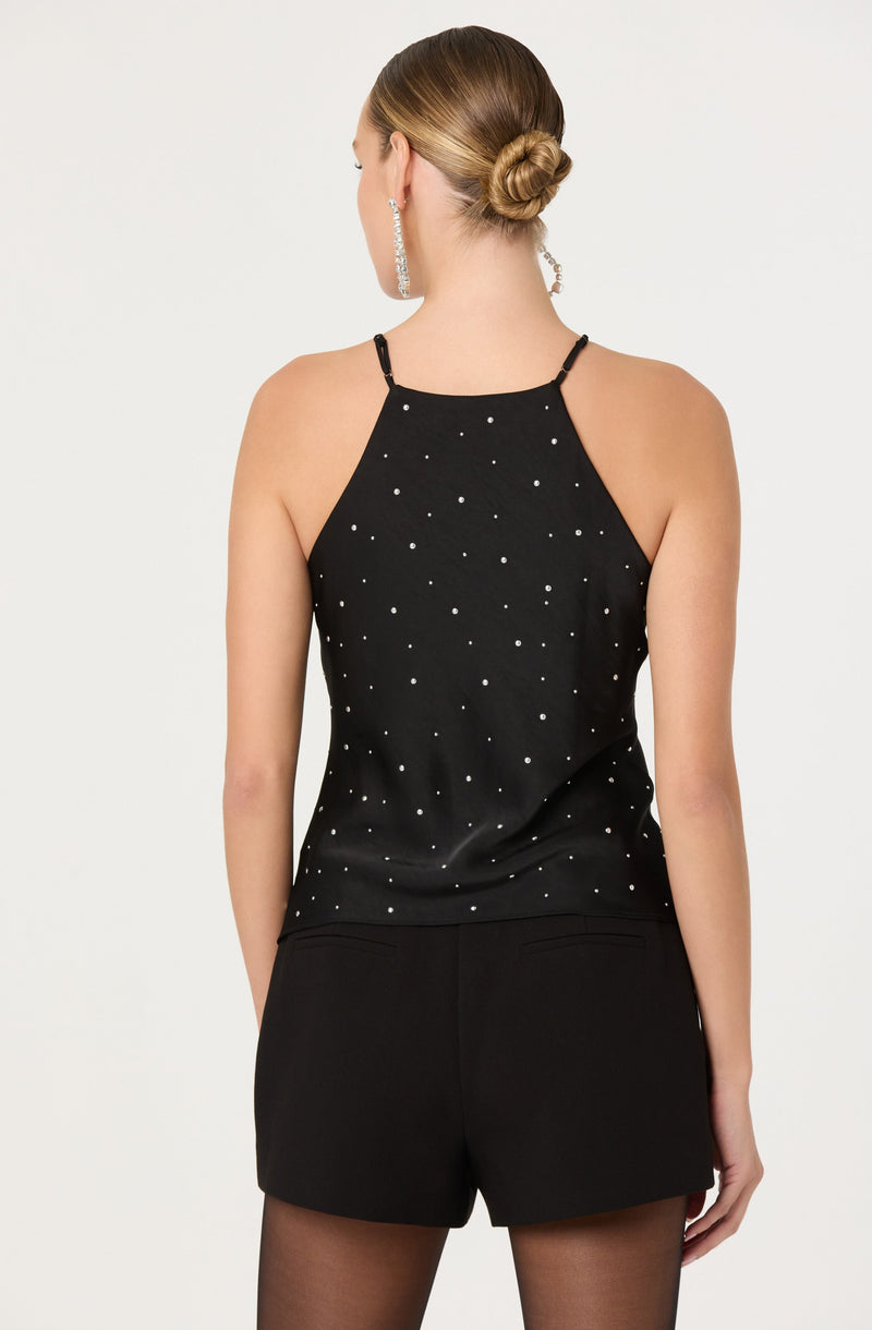 A woman is wearing a black sleeveless top with thin straps and a high neckline, decorated with small white polka dots. She is also dressed in black shorts, and she has her hair styled in a neat bun. She is accessorized with large dangling earrings.
