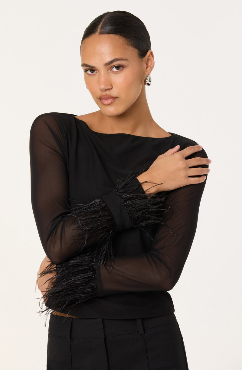 A woman is wearing a black long-sleeve top with sheer fabric, adorned with feathered details at the cuffs. She has paired it with black pants.