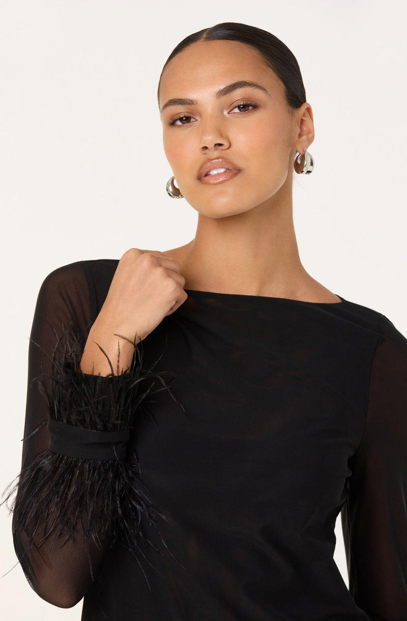 A woman is wearing a black long-sleeve top with sheer sleeves and feathered cuffs. She has her hair slicked back and is accessorized with large silver hoop earrings.