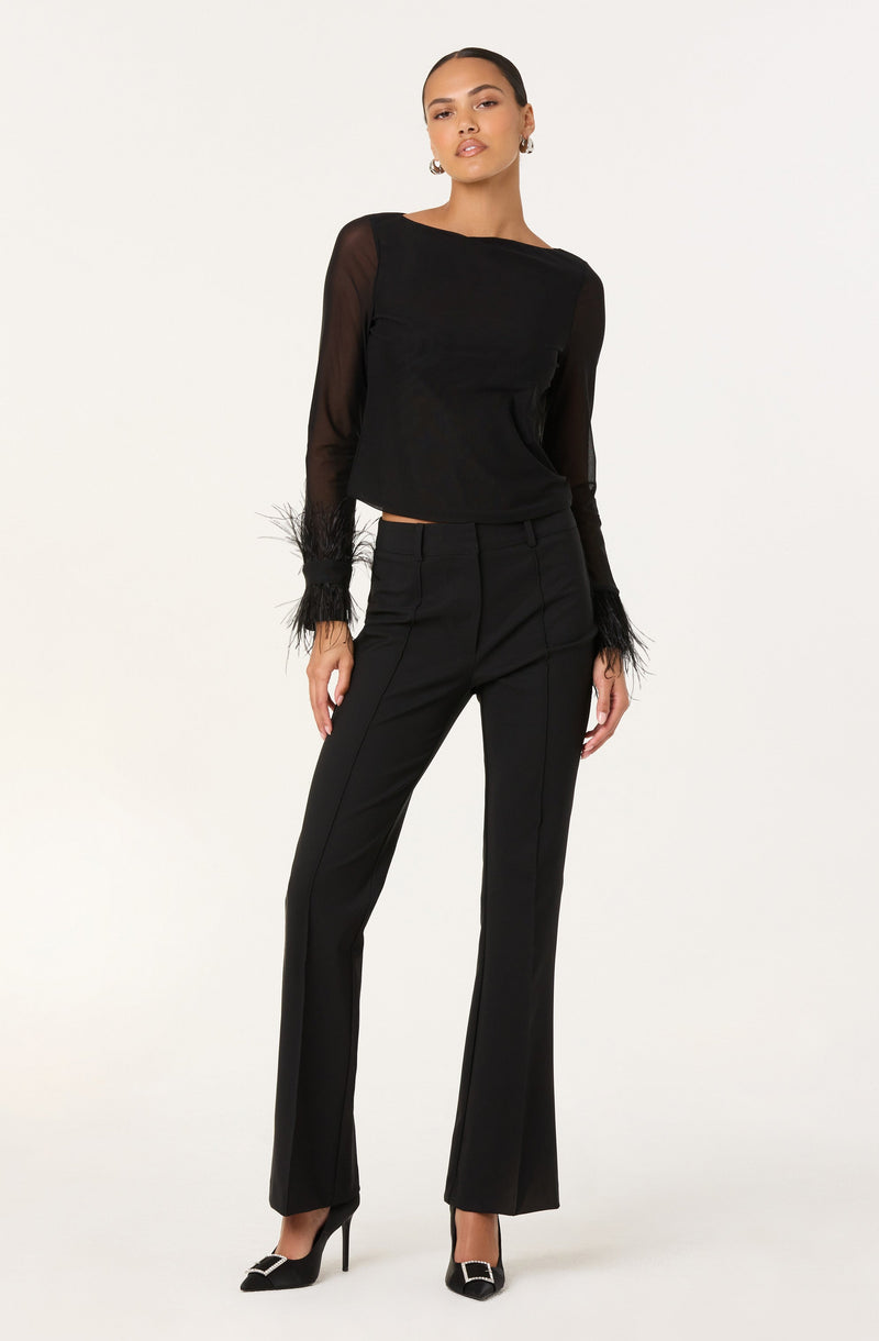 A woman is wearing a black long-sleeve top made of sheer fabric with feathered cuffs. She pairs it with high-waisted black trousers that have a straight-leg fit. She completes the outfit with black pointed-toe high heels featuring a decorative buckle on the toe.